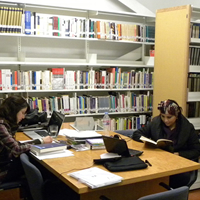 2 mujeres leyendo en una habitación, al fondo se pueden ver repisas repletas de libros